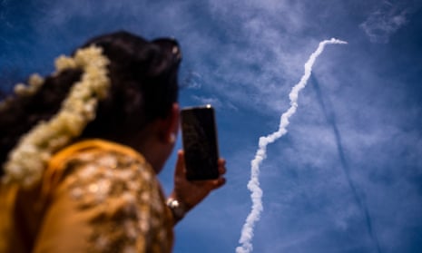 A rear-view image of woman holding up a smarthphone pointed at the vapour trail of the rocket launcher as it ascends through the sky.