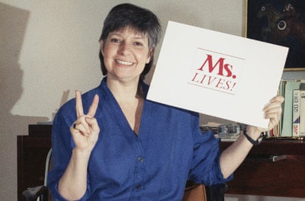 A smiling woman showing the V/victory sign with one hand while holding a sign that says "Ms Lives" with the other.