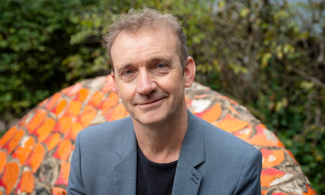 Un hombre de mediana edad con cabello corto y rizado, vistiendo una chaqueta gris y una camiseta negra, sonríe levemente mientras posa al aire libre. De fondo se puede apreciar un mosaico colorido con tonos naranjas y un entorno natural con vegetación difusa. Un hombre de mediana edad con cabello corto y rizado, vistiendo una chaqueta gris y una camiseta negra, sonríe levemente mientras posa al aire libre. De fondo se puede apreciar un mosaico colorido con tonos naranjas y un entorno natural con vegetación difusa.