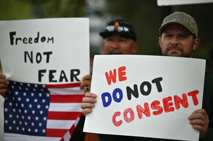 Two men hold protest signs and an American flag. One reads "Freedom not fear" and the other: "We do not consent".