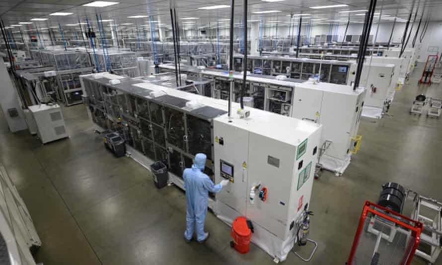 View of a large clean factory with rows of modern white equipment. One person is visible in full blue overalls as would be worn in a clean-room