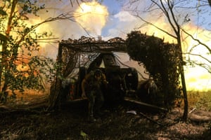 A soldier in camouflage turns away from a huge gun which has just been fired; it is emitting a huge wave of yellow flames. The gun is part-covered by camouflage netting and placed between two tiny, bare trees
