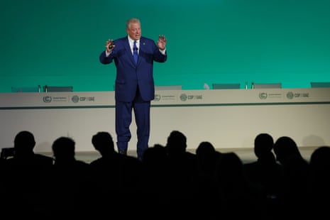 A man in a blue suit speaking and gesturing on a stage in front of a crowd in silhouette.