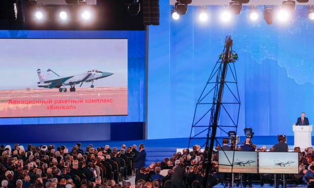 The video screen shows the Kinzhal air missile system as Russia’s President Vladimir Putin (R) delivers his annual address