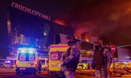 law enforcement holding guns outside building with sign saying crocus city hall with fire in the air behind them