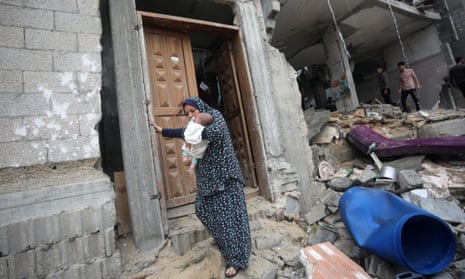 A woman in an abbaya picks her way through wreckage on a bombed street while holding a small baby
