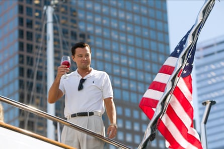 A man in grey chinos and a white polo shirt standing on a yacht in front of a skyscaper holding up a glass of rose wine with a US flag to his right