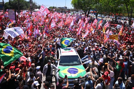 Lula holds a Brazil flag while standing atop a car. He is surrounded by a sea of supporters, many waving national flags and banners