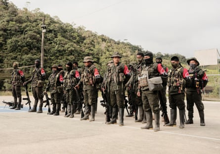 A group of people in green military uniforms with red armbands stands on a concrete court.