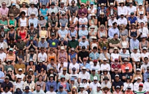 People sit shoulder to shoulder in tiered rows with not an empty seat in sight. Out of focus in the foreground, a tennis ball appears to hang in the air