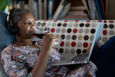 A middle-aged black lady wearing headphones sits sipping wine from a large glass watching a video on a laptop, reclining on a polka-dot-patterned sofa.