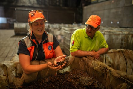 Lucy Harler, holds a sample of compost over a large bin of it in a shed, with Julius Joel standing beside her