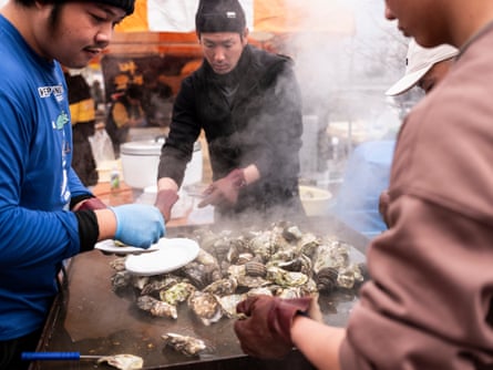 Three men stand around a steaming grill plate on which oysters are cooking