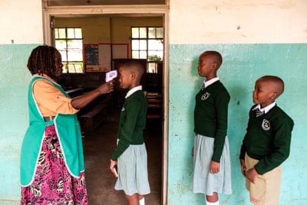 A nurse places a thermometer to the head of a child in school uniform, with two other children lining up behind