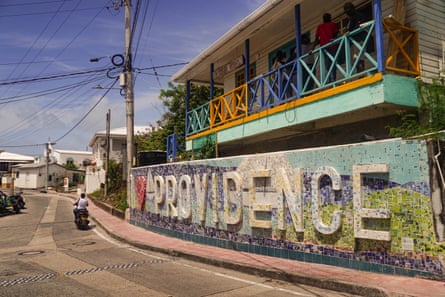 A street on Providence Island has a long tiled wall with letters reading I heart Providence in front of a building with a brightly coloured blue, gold and green painted veranda.