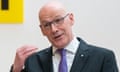 John Swinney gestures while speaking at a podium with a 'John Swinney' sign on the wall behind him