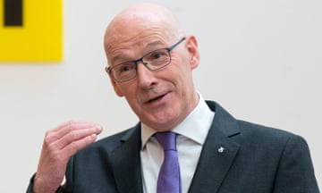 John Swinney gestures while speaking at a podium with a 'John Swinney' sign on the wall behind him
