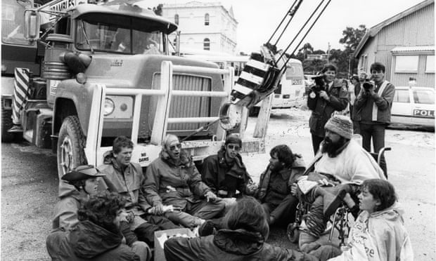 A black and white photo of a group of people sitting on the ground in a circle in front of a truck carrying a crane on it
