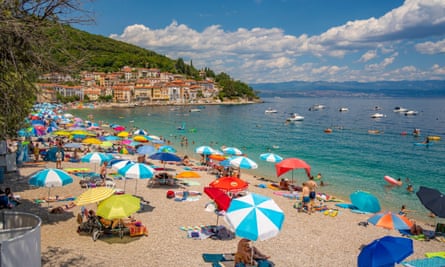 A sandy beach dotted with sun umbrellas in Croatia, with houses on the curved coastline, a cloudy sky and people, boats and rubber rafts in the clear water