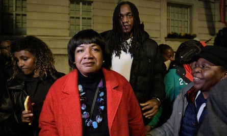Diane Abbott is pictured in front of Hackney town hall at an anti-racism rally. She is wearing a bright red coat and black top with a chunky blue and turquoise necklace, and is standing among a crowd.