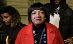 Diane Abbott in front of Hackney town hall at a rally held in her support; she is face-on to the camera looking resolute. It is dark and she wears a bright red coat.