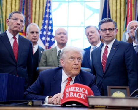 Donald Trump, surrounded by Republican lawmakers, with a red baseball cap reading 'America is Back!' on the desk