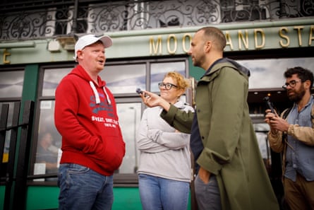 Polanski holds a microphone up to a man in a bright red hoodie and white baseball cap outside a pub