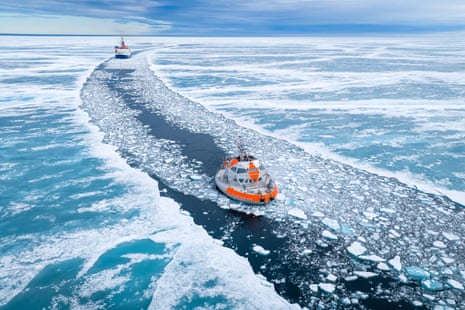 A drone shot from above of a small oval ship in a channel surrounded by sheet and pack ice with a more conventional ship in the distance