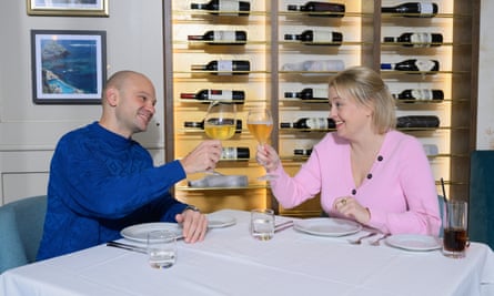 A bald man wearing a bright blue patterned jumper, and a blond woman wearing a pink cardigan, sitting at a restaurant table raising a toast