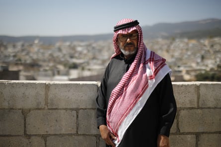 A middle-aged Arab man in a black thobe and red and white keffiyeh stands by a wall on a hill overlooking densely packed homes.
