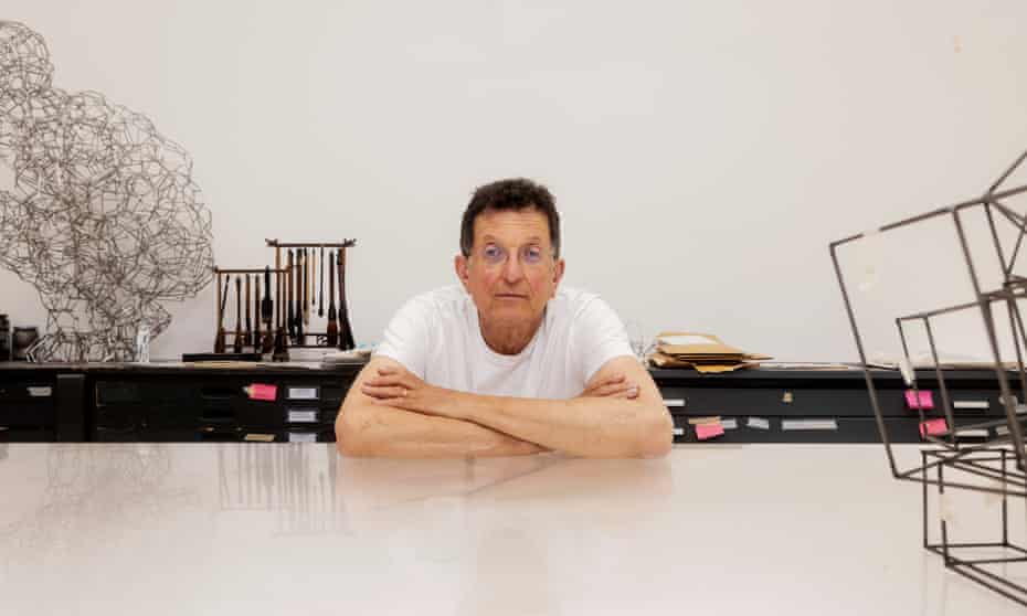 Antony Gormley posing with his arms folded, leaning on a large pale table with artworks in view in the background