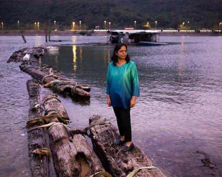 A woman in a green and blue top and dark trousers stands on a makeshift bridge fashioned from tree trunks within Hong Kong harbour