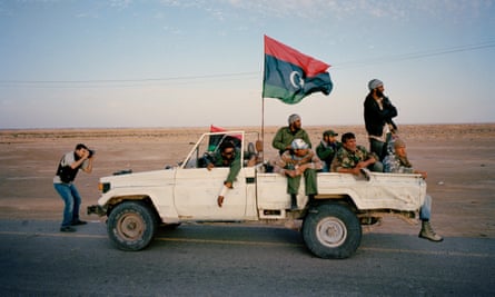 Fighters in a pick-up truck displaying a Libyan flag on an empty road, while a photographer takes their picture.