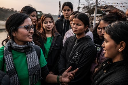 A woman talks as a group of people listen. They are outside and a body of water can be seen behind them