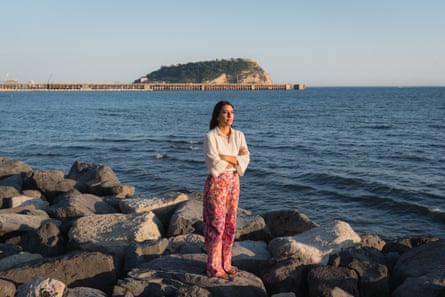 A woman wearing a white blouse and pink, flowery trousers stands with the arms folded on rocks at the water’s edge, looking along the coastline