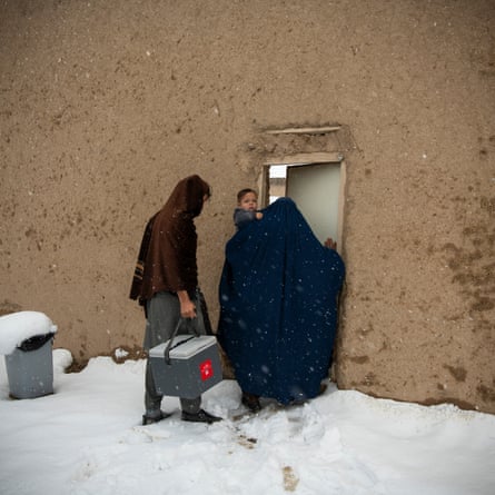 A man carrying a box by a strap follows a woman in a burqa holding a young child as she walks through the snow into a doorway