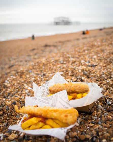 Two fish and chips on the beach on a pebbled beach in Brighton, with the West Pier in the distance.