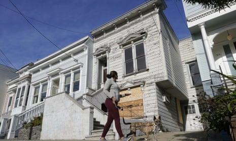 A woman walks a dog down a street in front of a dilapidated gray Victorian home.