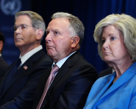 Two older men in dark suits and a woman in a blue outfit look to the left of the camera frame.