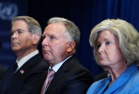 Two older men in dark suits and a woman in a blue outfit look to the left of the camera frame.