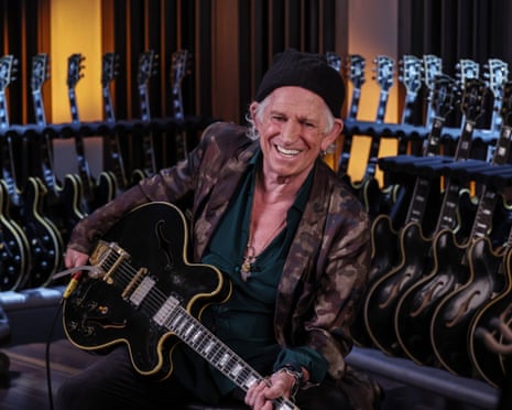 Keith Richards holding a guitar, with racks of guitars behind him