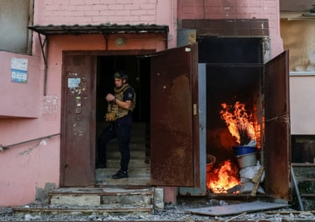 A man in a helmet and bulky waistcoat with first-aid kit stands in a doorway. In a cupboard to the side large flames are rising among buckets and other items. There is debris strewn around the entrance.