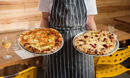A cropped photo shows someone wearing a striped chefs apron holding a pizza in each hand
