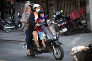 Woman rides a moped with a young child in front and an older child behind. The young child is holding a toy gun.