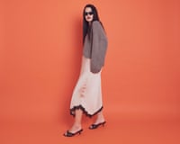 A young woman with long dark hair and red lipstick, wearing a baggy light brown jumper, a cream silky skirt with black lace trim, and leopard-print heeled sandals, standing against a deep orange background