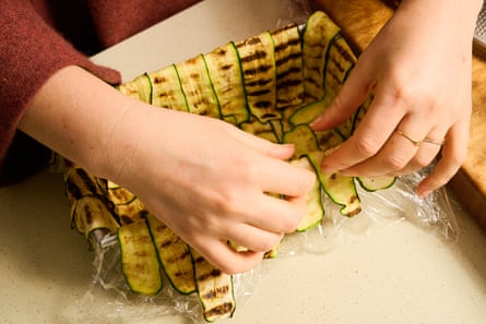 Two hands arranging grilled zucchini slices in a rectangular baking tin.