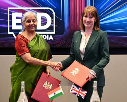 Rachel Reeves shakes hands with Nirmala Sitharaman in a conference room; they hold tiny Indian and union jack flags and red folders; Reeves wears a dark green trouser suit and Sitharaman wears a dark orange top with lighter green sari draped around her.
