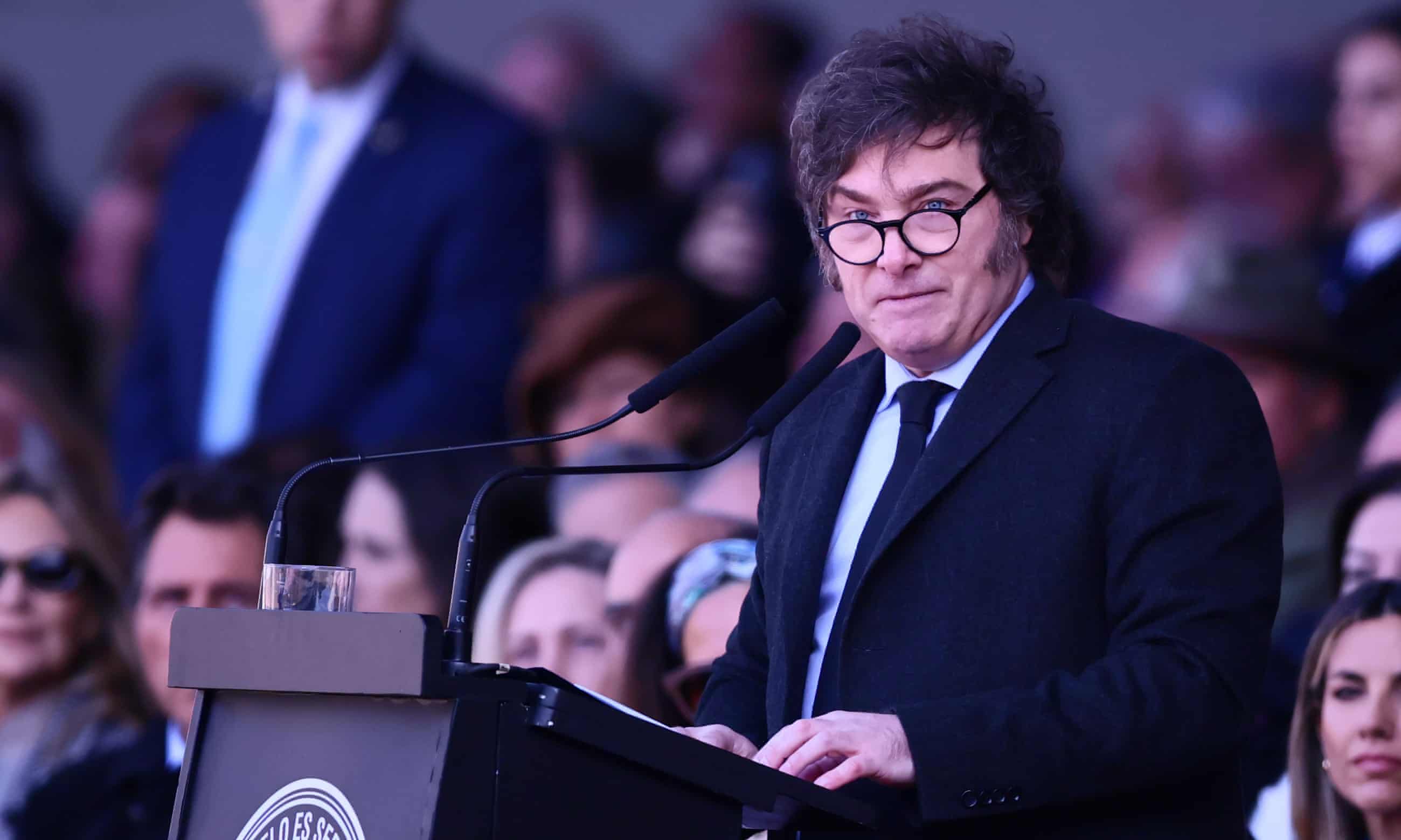 El presidente de Argentina, Javier Milei, hablando en un podio durante un evento, vestido con traje oscuro y corbata, y usando gafas. Detrás de él, se ven varias personas en el público desenfocadas.