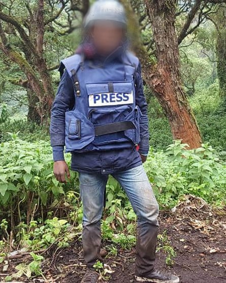 A man in muddy jeans and a press vest and helmet stands in an area of jungle.