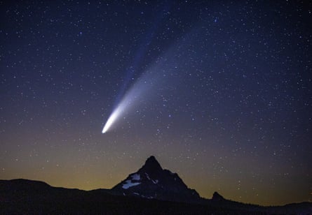 A comet and its tails streaking across a starry sky, above a mountain.